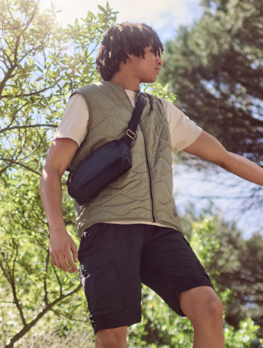 Ein junger Mann mit lockigen Haaren steht in einer natürlichen Umgebung mit Bäumen und blauem Himmel. Er trägt eine gesteppte Weste in Olivgrün, ein beiges T-Shirt, schwarze Shorts und eine schwarze Umhängetasche. Seine Körperhaltung wirkt dynamisch