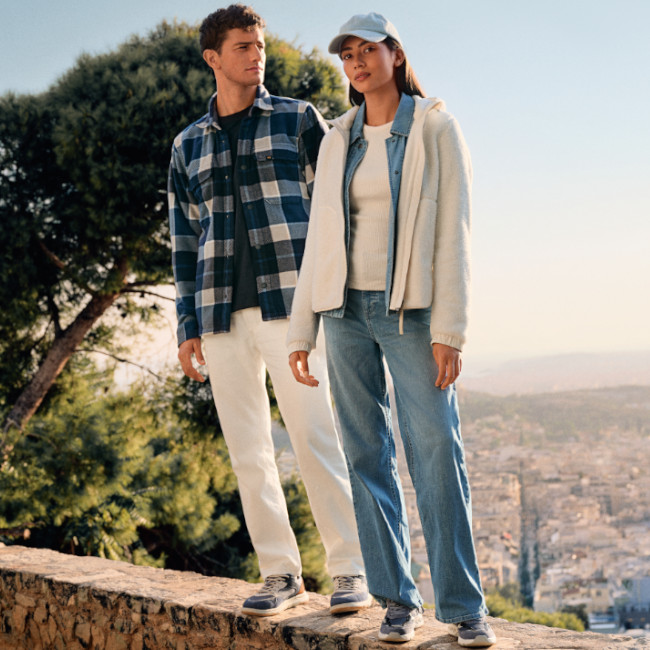 Ein Mann und eine Frau stehen in tropischer Umgebung. Der Mann tr&auml;gt ein hellblaues Hemd mit schwarzer Weste, die Frau eine gemusterte Bluse und dunkle Hose. Beide blicken in verschiedene Richtungen.