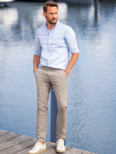 Ein Mann mit kurzem Bart und einem gepflegten Look steht entspannt auf einem Holzsteg an einem Gewässer. Er trägt ein hellblaues, langärmeliges Hemd mit hochgekrempelten Ärmeln, beige Chinohosen und weiße Sneaker. 