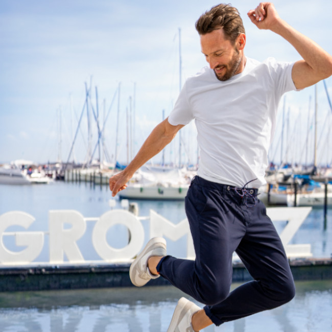 Ein Mann mit weißem Shirt und blauer Chinohose springt hoch. Im Hinterfung ist ein Hafen mit Segelboten zu sehen. 
