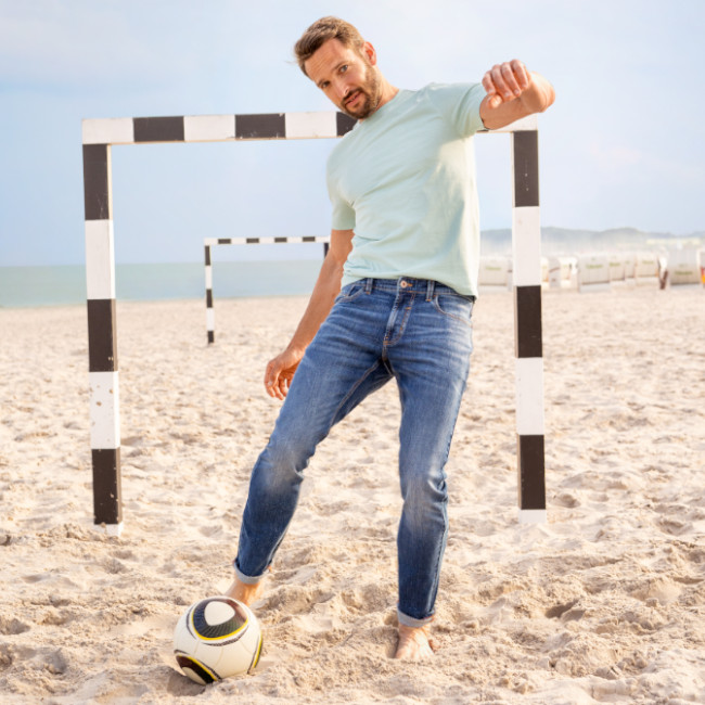 ein Mann spielt am Stand Fussball. Im Hintergrund ist ein Tor zu sehen. Er trägt eine Jeans und ein grünes Shirt.