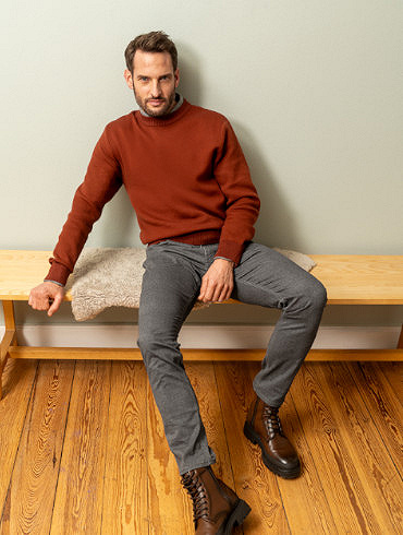 Ein Mann mit kurzem Bart und einem gepflegten Look steht entspannt auf einem Holzsteg an einem Gewässer. Er trägt ein hellblaues, langärmeliges Hemd mit hochgekrempelten Ärmeln, beige Chinohosen und weiße Sneaker. 