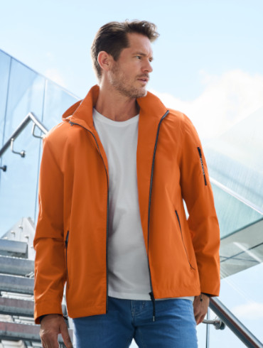 Ein Mann mit markanten Gesichtszügen steht auf einer modernen Treppe mit Glasgeländer. Er trägt eine auffällige orangefarbene Jacke mit einem hochgeklappten Kragen, ein weißes T-Shirt und blaue Jeans. Sein Blick ist entschlossen zur Seite gerichtet.