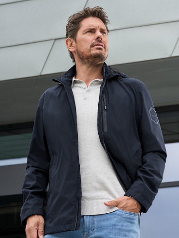 Ein Mann mit markanten Gesichtsz&uuml;gen steht auf einer modernen Treppe mit Glasgel&auml;nder. Er tr&auml;gt eine dunkelblaue Jacke mit einem hochgeklappten Kragen, ein wei&szlig;es T-Shirt und blaue Jeans. Sein Blick ist entschlossen zur Seite gerichtet.